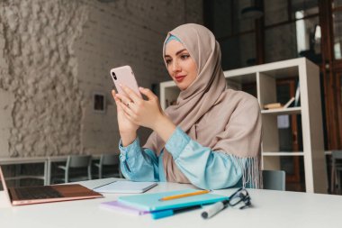 Genç, modern, tesettürlü Müslüman bir kadın ofis odasında dizüstü bilgisayarla çalışıyor, internetten eğitim görüyor, uzaktan kumandalı, akıllı telefon kullanıyor.