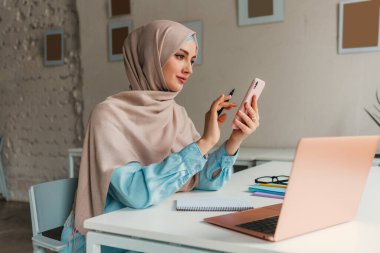 Genç, modern, tesettürlü Müslüman bir kadın ofis odasında dizüstü bilgisayarla çalışıyor, internetten eğitim görüyor, uzaktan kumandalı, akıllı telefon kullanıyor.