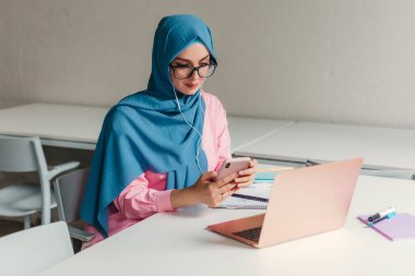 Genç, modern, tesettürlü Müslüman bir kadın ofis odasında dizüstü bilgisayarla çalışıyor. İnternette eğitim görüyor, uzaktan kumandalı çalışıyor. Akıllı telefon ve kulaklık kullanarak online konferans sohbeti yapıyor.