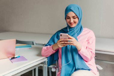Genç, modern, tesettürlü Müslüman bir kadın ofis odasında dizüstü bilgisayarla çalışıyor. İnternette eğitim görüyor, uzaktan kumandalı çalışıyor. Akıllı telefon ve kulaklık kullanarak online konferans sohbeti yapıyor.