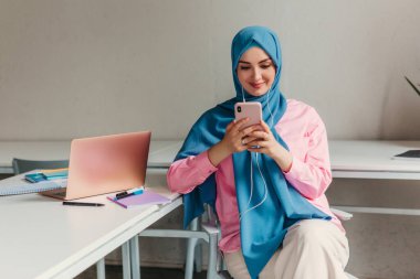 Genç, modern, tesettürlü Müslüman bir kadın ofis odasında dizüstü bilgisayarla çalışıyor. İnternette eğitim görüyor, uzaktan kumandalı çalışıyor. Akıllı telefon ve kulaklık kullanarak online konferans sohbeti yapıyor.