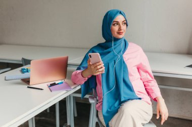 Genç, modern, tesettürlü Müslüman bir kadın ofis odasında dizüstü bilgisayarla çalışıyor. İnternette eğitim görüyor, uzaktan kumandalı çalışıyor. Akıllı telefon ve kulaklık kullanarak online konferans sohbeti yapıyor.