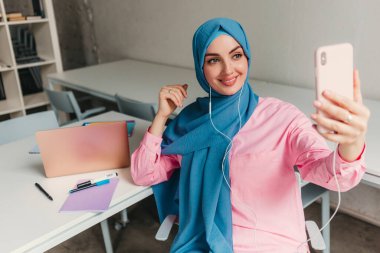 Genç, modern, tesettürlü Müslüman bir kadın ofis odasında dizüstü bilgisayarla çalışıyor. İnternette eğitim görüyor, uzaktan kumandalı çalışıyor. Akıllı telefon ve kulaklık kullanarak online konferans sohbeti yapıyor.