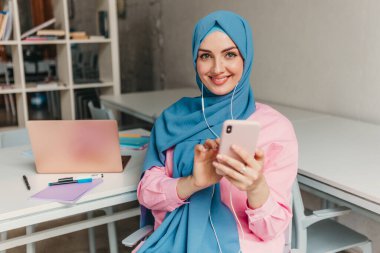 Genç, modern, tesettürlü Müslüman bir kadın ofis odasında dizüstü bilgisayarla çalışıyor. İnternette eğitim görüyor, uzaktan kumandalı çalışıyor. Akıllı telefon ve kulaklık kullanarak online konferans sohbeti yapıyor.
