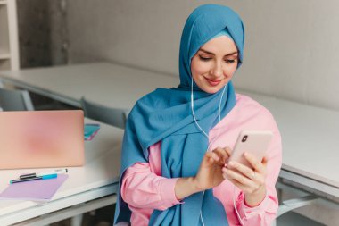 Genç, modern, tesettürlü Müslüman bir kadın ofis odasında dizüstü bilgisayarla çalışıyor. İnternette eğitim görüyor, uzaktan kumandalı çalışıyor. Akıllı telefon ve kulaklık kullanarak online konferans sohbeti yapıyor.