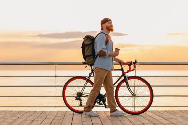 Sırt çantalı, kot gömlekli ve şapkalı yakışıklı hippi tarzı sakallı bir adam. Gün doğumunda deniz kenarında bisiklet giyiyor. Kahve içiyor.