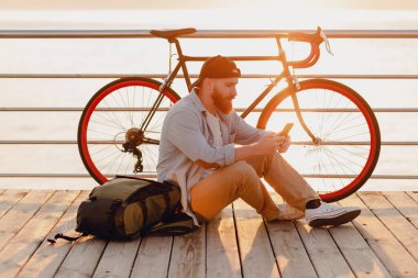 Sırt çantalı yakışıklı hippi tarzı sakallı adam elinde akıllı bir telefon, kot gömlek giyiyor ve sabahleyin bisiklet takıyor. Deniz kenarında sağlıklı, aktif bir hayat tarzı gezgini.