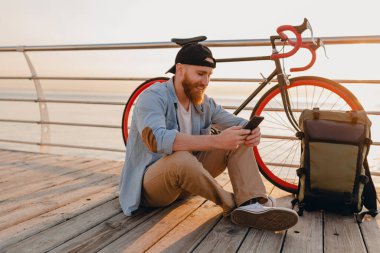 Sırt çantalı yakışıklı hippi tarzı sakallı adam elinde akıllı bir telefon, kot gömlek giyiyor ve sabahleyin bisiklet takıyor. Deniz kenarında sağlıklı, aktif bir hayat tarzı gezgini.