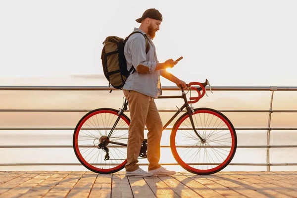 handsome hipster style bearded man traveling with backpack on bicycle using phone in morning sunrise by the sea, healthy active lifestyle