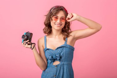 Çekici, gülümseyen bir kadın kot elbise ve güneş gözlükleriyle fotoğraf çekiyor. Pembe arka planda izole edilmiş, tatilde gezgin, yaz modası tarzı.