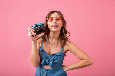 Komik gülümseyen duygusal yüz ifadesi olan çekici bir kadın kot elbiseli klasik kamera ve pembe arka planda izole edilmiş güneş gözlüklü, tatilde gezgin, yaz moda turisti
