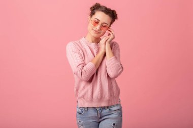 Genç, güzel, romantik bir kadın ellerini yüzüne koyup pembe süveter ve pembe gözlüklerle pembe stüdyo arka planında hayal kuruyor.