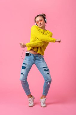 Sarı kazaklı çekici, neşeli bir kadın kulaklıkla müzik dinliyor. Hipster tarzı kıyafetler giyiyor. Pembe stüdyo geçmişinde izole edilmiş, eğleniyor.