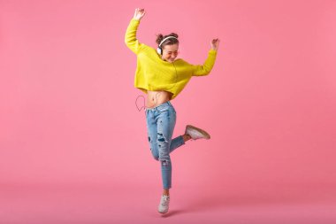 Çekici, mutlu, komik bir kadın kulaklıkla müzik dinliyor. Hipster renkli kıyafetler giyiyor. Pembe stüdyo arka planında izole edilmiş, sarı kazak ve güneş gözlüğü takıyor, eğleniyor.