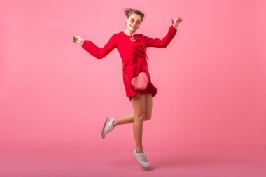 Çekici, mutlu, modaya uygun, kırmızı elbiseli, pembe stüdyo arka planında dans eden, yaz moda trendi, romantik ruh hali flörtçü kız.