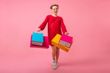 Kırmızı modaya uygun elbiseler içinde, pembe stüdyo arka planında renkli alışveriş çantaları taşıyan neşeli, mutlu, şık bir kadın.