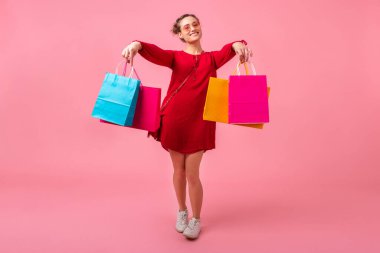 Çekici, mutlu, stil düşkünü, kırmızı modaya uygun elbiseler içinde alışveriş koliklerinin pembe stüdyo arka planında renkli alışveriş çantaları taşıması, indirimli satış heyecanı, moda trendi