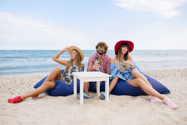 young hipster company of friends on vacation sitting at beach on bean bags, drinking mojito cocktail, happy, smiling, positive, summer style, fashion trend, spending good time together, party mood