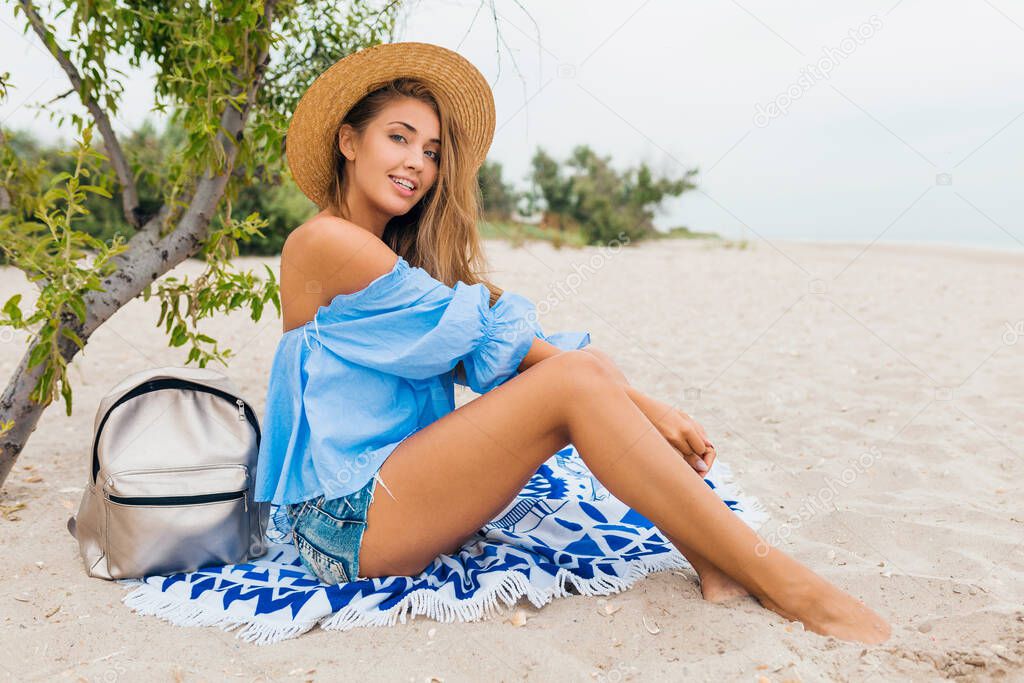 stylish beautiful smiling woman sitting on sand with skinny legs on summer vacation on tropical beach wearing straw hat, silver backpack