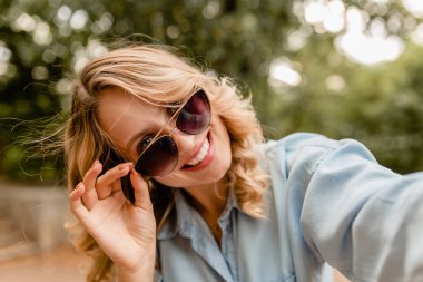 Çekici, sarışın, gülümseyen bir kadın yaz kıyafetleriyle parkta yürüyor. Mavi gömlekli, telefonda selfie çeken, zarif güneş gözlüklü, sokak modası tarzında.