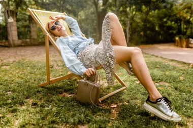 Çekici, sarışın, mutlu bir kadın güverte sandalyesinde oturuyor. Mavi gömlekli, gümüş rengi spor ayakkabılı, şık güneş gözlüklü ve çantalı, sokak modası tarzı ayakkabılı.