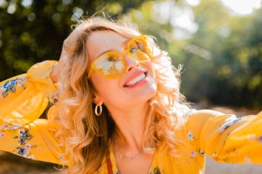 Sarı bluz giyen, güneş gözlüğü takan, gülümseyen güzel sarışın beyaz dişli kadının portresi selen fotoğraf, renkli yaz modası, parlak bahar güneşli bir gün, pozitif mutlu bir ruh hali.