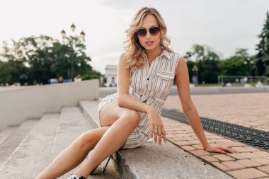 Genç, çekici, şık, sarışın, gülümseyen kadın yazın caddede oturuyor. Moda tarzı beyaz elbise, güneş gözlüğü takıyor, seksi ve zarif bir tarzı var.