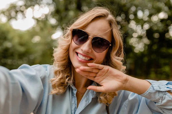 attractive blond smiling white teeth woman walking in park in summer outfit blue shirt taking selfie photo on phone, wearing elegant sunglasses, street fashion style