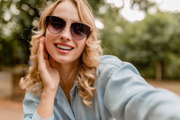 魅力的な金髪の笑顔の女性が夏の服装で公園を歩いています携帯電話で自撮り写真を撮る青いシャツ エレガントなサングラスを身に着けて ストリートファッションスタイル — ストック写真