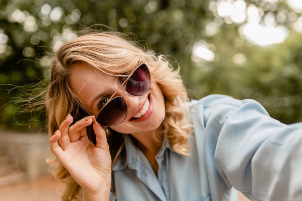 魅力的な金髪の笑顔の女性が夏の服装で公園を歩いています携帯電話で自撮り写真を撮る青いシャツ エレガントなサングラスを身に着けて ストリートファッションスタイル — ストック写真