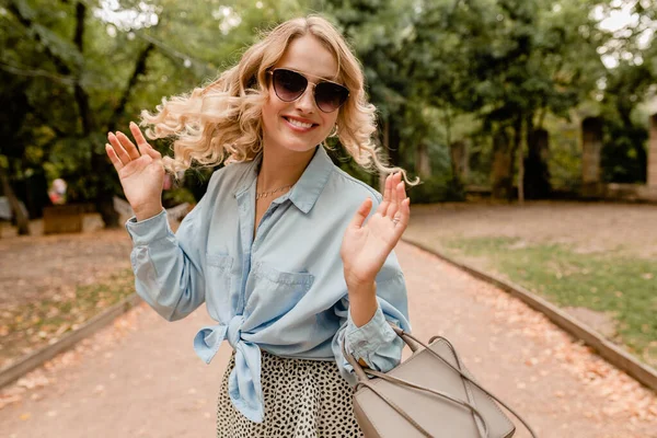 attractive blond smiling candid woman walking in park in summer outfit blue shirt wearing elegant sunglasses and purse, street fashion style, happy mood, waving curly hair