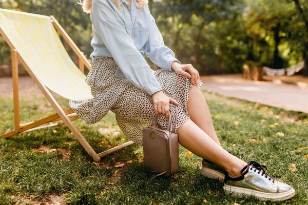 close-up legs in silver sneakers shoes, fashion autumn trend footwear, attractive woman sitting in deck chair in stylish outfit blue shirt and skirt, holding purse, street style accessories