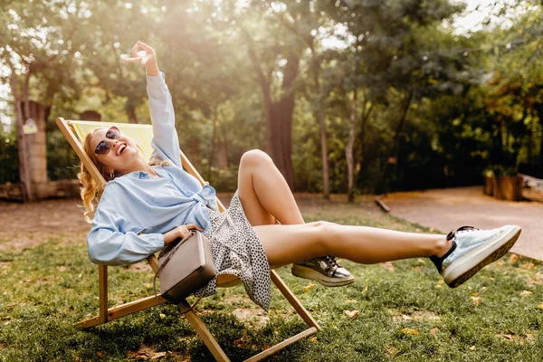夏の衣装でデッキチェアでリラックスして座って魅力的なブロンドの幸せな女性ブルーシャツ シルバースニーカー エレガントなサングラスと財布 ストリートファッションスタイルの履物を身に着けている — ストック写真