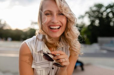 Yakından çekilmiş genç, şık sarışın kadın portresi. Yazın moda elbisesi içinde. Güneş gözlüğü takıyor, samimi bir gülümsemeyle gülüyor.