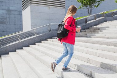Pembe montlu genç hippi kadın, sırt çantası ve kahveyle merdivenlerde yürüyen kot pantolon kulaklıkla müzik dinliyor, büyük şehir tarzı trend, güneş gözlüğü takıyor.