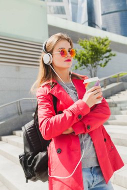 Pembe ceketli genç hippi kadın, sırt çantalı kot pantolon ve kahve kulaklıkla müzik dinliyor, büyük şehir tarzı trend, güneş gözlüğü takıyor.
