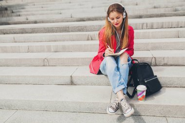 Pembe ceketli genç hippi kadın, kot pantolonlu, sırt çantalı ve kahveli kulaklıklı müzik dinleyen, büyük şehir tarzı trend, notlar alan öğrenci