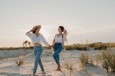 Güzel iki genç kadın gün batımında kumsalda eğleniyor. İkili cinsiyet ayrımcılığı yapmayan eşcinsel lezbiyen aşk ilişkisi. Kot pantolon giyen boho yaz tatili tarzı.
