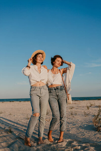 cool two young women having fun on the sunset beach, queer non-binary gender identity, gay lesbian love romance, boho summer vacation style wearing jeans