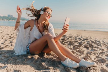 Genç, çekici, sarışın, gülümseyen bir kadın tatilde telefonla fotoğraf çekiyor. Plajda kulaklıkla oturmuş müzik dinliyor.