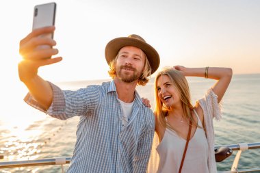 Genç, çekici, güler yüzlü, mutlu bir adam ve kadın bisikletle seyahat ediyor. Telefon kamerasıyla selfie çekiyor. Deniz kenarında romantik bir çift gün batımında.