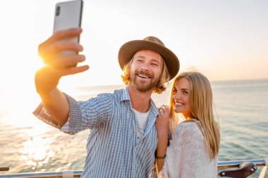 Genç, çekici, güler yüzlü, mutlu bir adam ve kadın bisikletle seyahat ediyor. Telefon kamerasıyla selfie çekiyor. Deniz kenarında romantik bir çift gün batımında.
