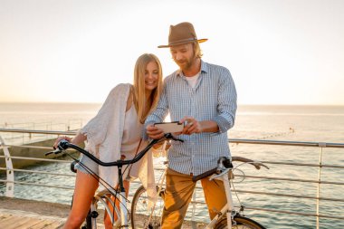 Genç, çekici, güler yüzlü, mutlu erkek ve kadın bisikletle seyahat ederken akıllı telefondan bakıyor, gün batımında deniz kenarında romantik bir çift, boho hipster kıyafeti, birlikte eğlenen arkadaşlar.