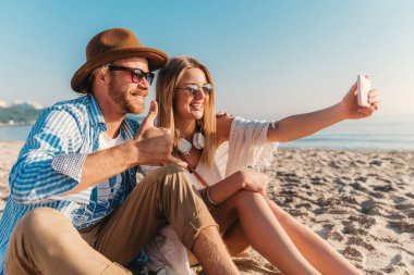Genç, çekici, güler yüzlü, güneş gözlüklü bir adam ve bir kadın kumsalda oturmuş telefon kamerasında selfie çekiyorlar. Gün batımında deniz kenarında romantik bir çift.