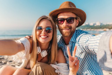 Genç, çekici, güler yüzlü, güneş gözlüklü bir adam ve bir kadın kumsalda oturmuş telefon kamerasında selfie çekiyorlar. Gün batımında deniz kenarında romantik bir çift.