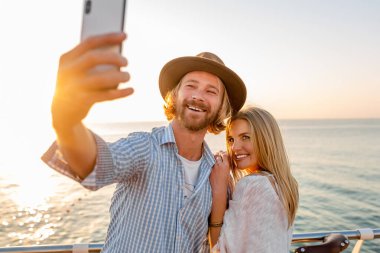 Genç, çekici, güler yüzlü, mutlu bir adam ve kadın bisikletle seyahat ediyor. Telefon kamerasıyla selfie çekiyor. Deniz kenarında romantik bir çift gün batımında.