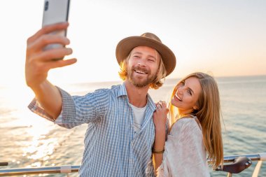Genç, çekici, güler yüzlü, mutlu bir adam ve kadın bisikletle seyahat ediyor. Telefon kamerasıyla selfie çekiyor. Deniz kenarında romantik bir çift gün batımında.