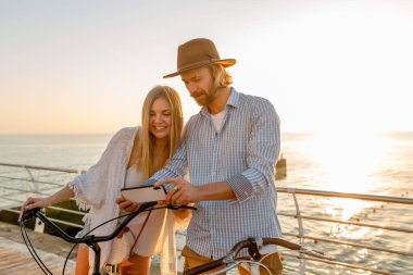 Genç, çekici, güler yüzlü, mutlu bir adam ve kadın bisiklete biniyor, akıllı telefon kullanıyor, romantik bir çift gün batımında deniz kenarında, boho hipster tarzı kıyafetler, arkadaşlar birlikte eğleniyor.