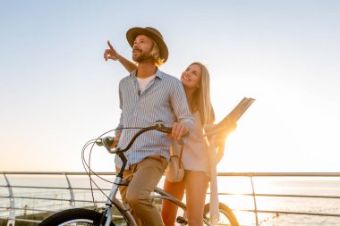 Yazın bisikletle seyahat eden çekici ve mutlu bir çift, Boho hipster tarzı giyinmiş bir kadın ve erkek birlikte eğleniyor, gezip fotoğraf çekiyor, parmakla işaret ediyor.