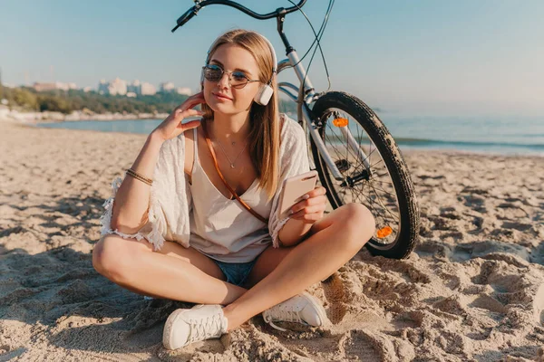スタイリッシュな若い魅力的なブロンドの笑顔の女性は自転車でビーチに座ってヘッドフォンで音楽を聞いてリラックス電話を使用して — ストック写真